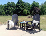 Two Krahn poly Adirondack chairs with a small table on a patio area with trees in the background.